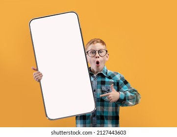 Wow, Great Offer. Excited Little School Boy In Eyeglasses Holding Big Cell In Hand Pointing At Phone Empty Screen Recommending App On Yellow Orange Studio Background. Smartphone Display Mock Up