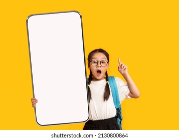 Wow, Great Offer. Excited Little Asian School Girl In Eyeglasses Holding Big Cell In Hand Pointing Finger Up Presenting Phone Empty Screen On Yellow Orange Studio Background. Smartphone Display Mockup