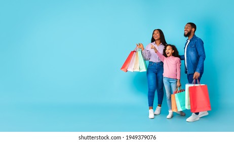 Wow, Great Offer. Full body length shot of African American family of three people holding shopping bags with new clothes or gifts, excited girl pointing aside at free copy space, blue studio wall