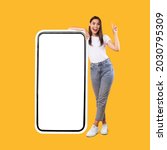 Wow. Happy Woman Leaning On Big Smartphone With Blank White Screen And Pointing Finger Up, Cheerful Lady Recommending New App Or Website, Standing On Yellow Background, Mock Up Image, Full Body Length