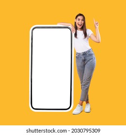 Wow. Happy Woman Leaning On Big Smartphone With Blank White Screen And Pointing Finger Up, Cheerful Lady Recommending New App Or Website, Standing On Yellow Background, Mock Up Image, Full Body Length - Shutterstock ID 2030795309