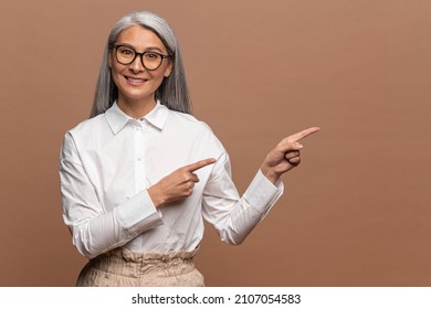 Wow, Look At Ads Here. Portrait Of Asian Elderly Woman Pointing Aside, Showing Blank Copy Space For Idea Presentation, Commercial Text. Indoor Studio Shot Isolated On Beige Background