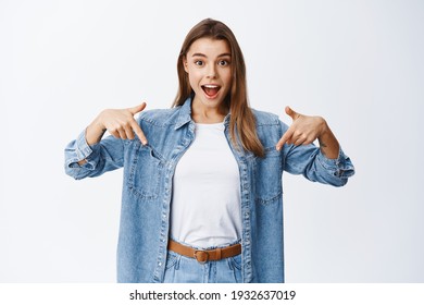 Wow look there. Excited blond young woman pointing fingers down with surprised face, showing awesome promo, demonstrating advertisement, white background