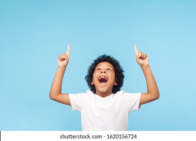 Wow, look up! Portrait of amazed little boy pointing up to empty place on blue background, expressing shock surprise with wide open mouth and showing copy space for advertisement. indoor studio shot
