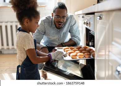 Wow, looks great! Excited caring millennial african dad or grown elder brother watching concentrated small black daughter or younger preteen sister taking pan with self baked tasty muffins out of oven