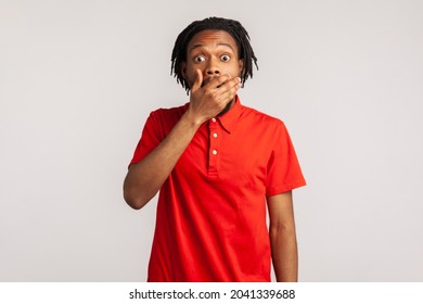Wow! Man wearing red casual T-shirt, covering mouth with hand and looking at camera with big eyes, scared surprised expression, shocked by sudden news. Indoor studio shot isolated on gray background.
