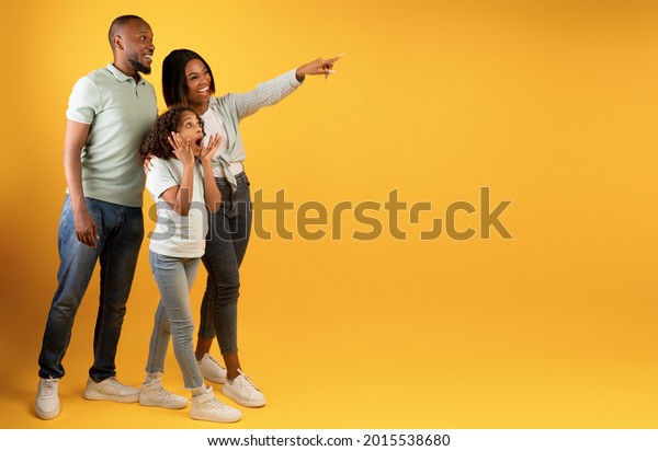 Wow offer. Full body length portrait of excited\
black family looking and pointing aside at copy space on yellow\
background, demonstrating free place for your advert, panorama,\
banner