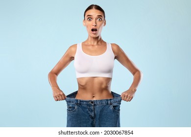 Wow. Portrait Of Shocked Suprised Young Woman Wearing Too Big Loose Jeans After Successful Weight Loss, Posing and Looking At Camera With Open Mouth In Amazement Standing Over Blue Studio Background