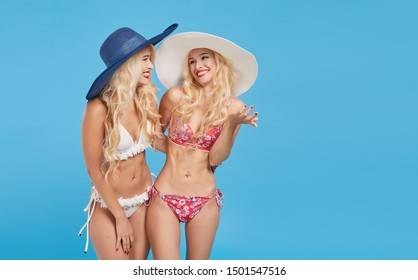 Wow And Shocked Content. Two Beautiful And Sexy Blond Tween Women In Bikini And Summer Hats On Blue Isolated Background. Studio Shot. Travel And Vacation Concept. Space For Text