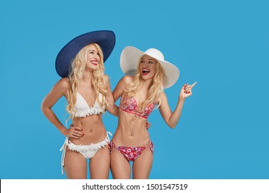 Wow And Shocked Content. Two Beautiful And Sexy Blond Tween Women In Bikini And Summer Hats On Blue Isolated Background. Studio Shot. Travel And Vacation Concept. Space For Text