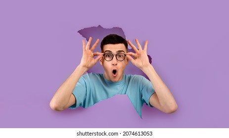 Wow, Unbelievable Offer. Portrait of shocked man in glasses with amazed expression looking through torn hole in violet paper. Panorama, banner, free copy empty space for advertising, purple background