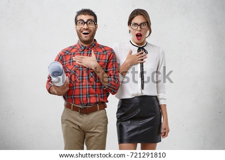 Wow! Wonderment and disbelief concept. Talented male architect looks funny as wears big spectales, holds rolled up paper or blueprint, stands next to girlfriend who helps to create new project work