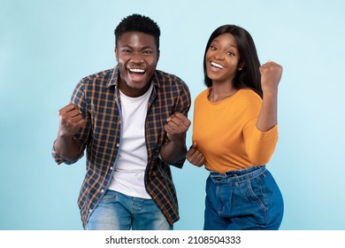 Wow, Yes. Portrait Of Overjoyed Young Black Man And Woman Cheering And Shaking Clenched Fists, Looking At Camera. Happy Couple Celebrating Win Posing Standing Isolated Over Blue Studio Background Wall