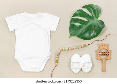 Wrap bodysuit for mockup with tropical monstera leaf on a pastel beige background in eco style. Teething beads, booties and cute wooden rattle for baby clothes mockup.
