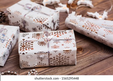 Wrapped In Festive Packaging Gift Boxes Tied With Silver Ribbon And  A Roll Of Wrapping Paper  On A Wooden Table. Preparing For Christmas.