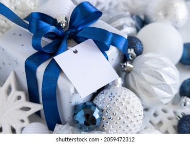 Wrapped Gift Box With A Blue Bow And Square Paper Gift Tag On A White Table With White And Silver Christmas Decorations Around Top View. Winter Composition With Blank Label Card, Mockup, Copy Space