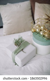 Wrapped Gift Box Decorated With Silk Ribbon And Fir Branch, Christmas Decorations In Basket On White Linen In Bedroom. Morning Gift Giving.