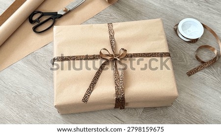 Wrapped gift box with leopard print ribbon and scissors on wooden surface, additional wrapping paper and tape visible in the background