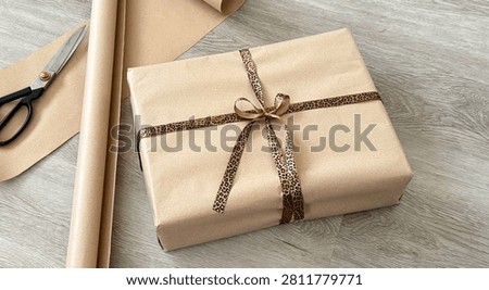Wrapped gift box with leopard print ribbon on wooden surface, surrounded by scissors and wrapping paper rolls in a well-lit indoor setting