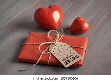 Wrapped gift with handmade craft cardboard paper tag tied up with cord and two red stone hearts on rustic wooden table. Text on the tag "Zero waste Christmas". Eco friendly Xmas gifts. - Powered by Shutterstock