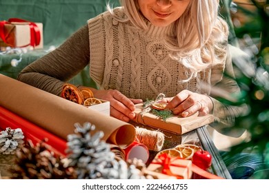 Wrapping christmas gifts. Blond woman wrapping presents in recycled card and decorated it with dried oranges and fir branches near the Christmas tree. Winter holiday celebration. - Powered by Shutterstock