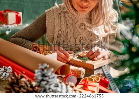 Wrapping christmas gifts. Blond woman wrapping presents in recycled card and decorated it with dried oranges and fir branches near the Christmas tree. Winter holiday celebration.
