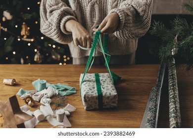 Wrapping christmas gifts. Hands in cozy sweater wrapping stylish present in festive wrapping paper with ribbons, vintage ornaments, bows on wooden table. Atmospheric winter holidays - Powered by Shutterstock