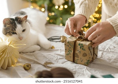 Wrapping stylish christmas gift, winter holidays preparation. Hands in cozy sweater decorating gift box and and cute cat helping on table on background of decorated christmas tree with lights - Powered by Shutterstock