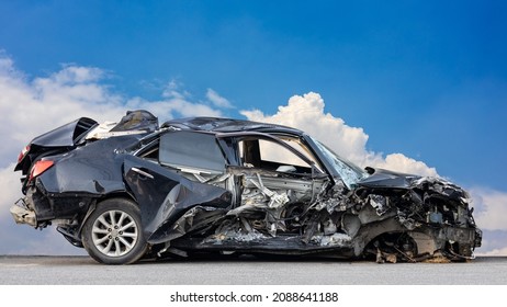 The wreckage of a black sedan smashed into wreckage until it turned into scrap in a fatal collision with another car with clouds and a blue sky in the background.
