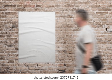 Wrinkled, blank A3 poster in white, adhered to a brick wall. A man walks beside. Ad or marketing campaign design promotion