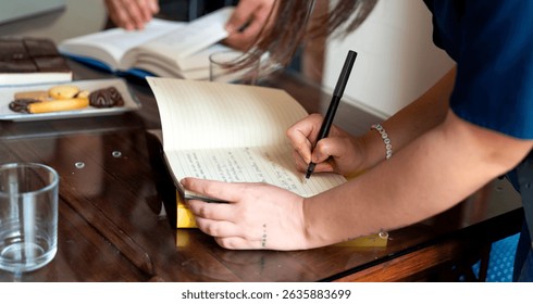 Writer taking notes in a notebook during a lively book club meeting, surrounded by cookies and refreshing glasses of water on the table