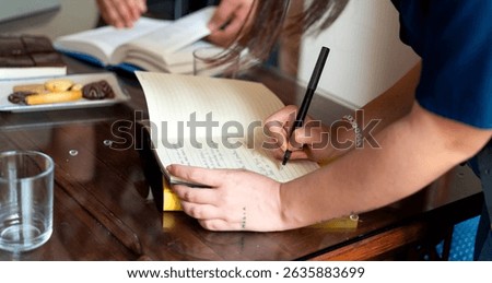 Writer taking notes in a notebook during a lively book club meeting, surrounded by cookies and refreshing glasses of water on the table