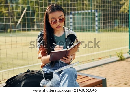 Writes in the notepad. Young asian woman is outdoors at daytime.