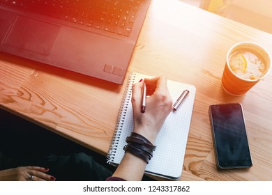 writes in small notebook, a hot cup of tea and a slice of orange. Workplace, pen and laptop, the working. coffee shop, phone, smartphone on the table, hands, study, warm color toning