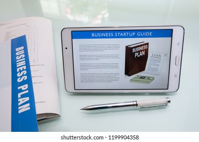 Writing A Business Plan Using A Business Startup Guide, Printed Collateral, Pen, And Tablet Or Mobile Device On A Reflective Glass Desk.