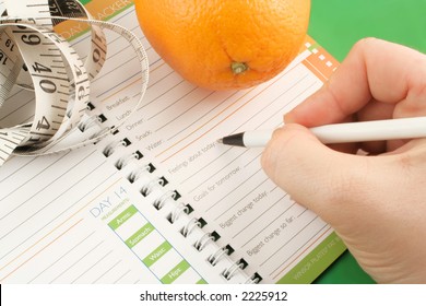 Writing In A Diet And Nutrition Journal With Orange And Tape Measure To The Side