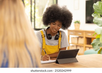 Writing in notepad, young woman using tablet with headphones in casual setting. Technology, notetaking, lifestyle, studying - Powered by Shutterstock