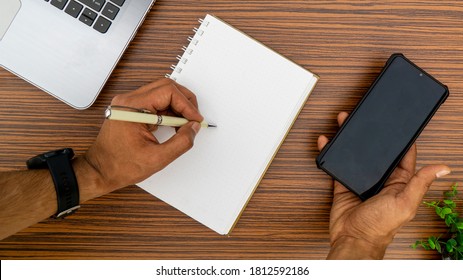 Writing on a notepad while holding a mobile phone working in an office environment. A lap top, a mobile, calculator and plant are also on display on this brown striped working table. - Shutterstock ID 1812592186