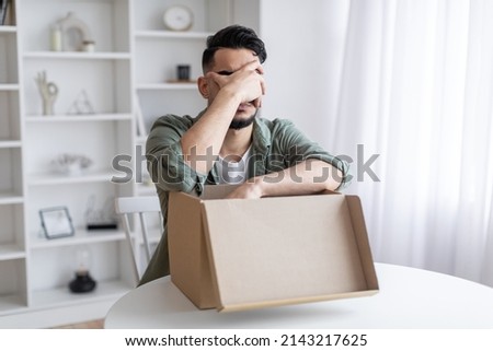 Wrong Item. Upset Arab Guy Opening Parcel And Making Facepalm Gesture While Sitting At Table At Home, Young Middle Eastern Man Feeling Sad After Unpacking Box With Delivery, Copy Space