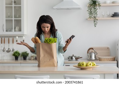 Wrong order, forgot to buy, mistake and bad delivery services. Disgruntled upset millennial african american woman looking in eco bag with groceries and hold smartphone, gesturing in kitchen interior
