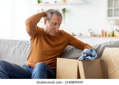 Wrong Parcel Concept. Shocked frustrated senior man unpacking box with shirt, unhappy with bad delivery service sitting on sofa at home in living room, grabbing head. Shipping mistake, free copy space