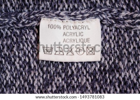 Wrong side of a blue sweater made of acryl, the composition is specified: 100% polyacryl (acrylic). Fabric composition white clothes label on blue texture background.