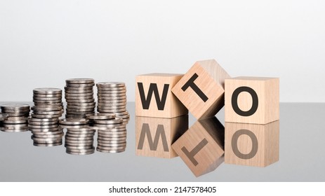 WTO - World Trade Organization - text is made up of letters on wooden cubes lying on a mirror surface, gray background. stacks with coins. inscription is reflected from the surface. selective focus.
