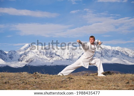 Wushu master in a white sports uniform training on the hill. Kungfu champion trains maritial arts in nature on background of snowy mountains.