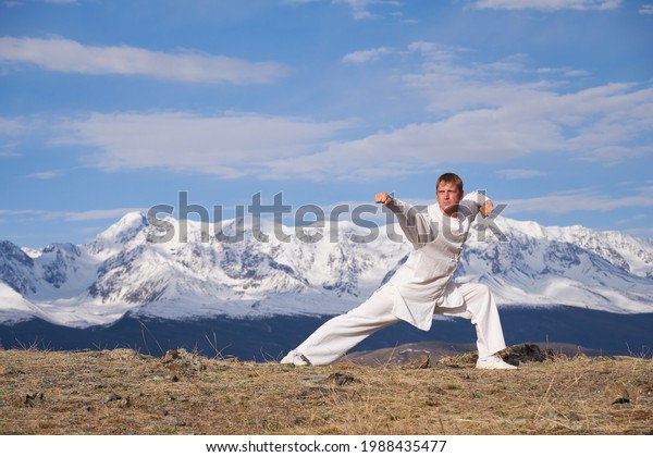Wushu master in a white sports uniform\
training on the hill. Kungfu champion trains maritial arts in\
nature on background of snowy\
mountains.
