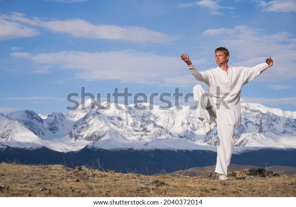 Wushu master in a white sports uniform\
training on the hill. Kungfu champion trains maritial arts in\
nature on background of snowy\
mountains.
