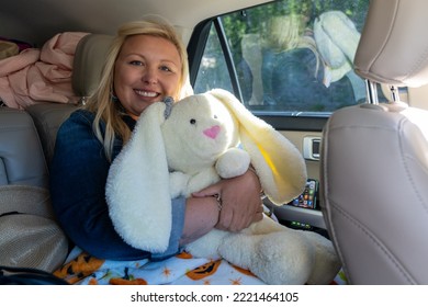 Wyoming, USA - July 20, 2022: Happy Blonde Woman Sits In The Backseat Of A Car, Holding And Hugging A Stuffed Animal Bunny