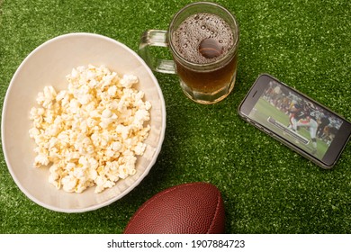 XALAPA, VERACRUZ, MEXICO- JANUARY 18, 2021: American Football Ball, Pop Corn And Cell Phone With American Football Game On Green Grass Background