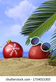 Xmas Summer Holidays Concept. Christmas Ornaments On A Sandy Beach With Palm Tree, Blue Sky And Sea Background. Vertical Photo