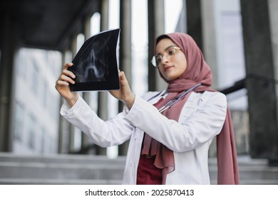 X-ray, Roentgen Image. Serious Thoughtful Muslim Woman Surgeon, Looking At X-Ray Radiography Of Patient's Head Skull. Female Arab Doctor Checking Xray Scan Outdoors. Selective Focus On X-ray Scan